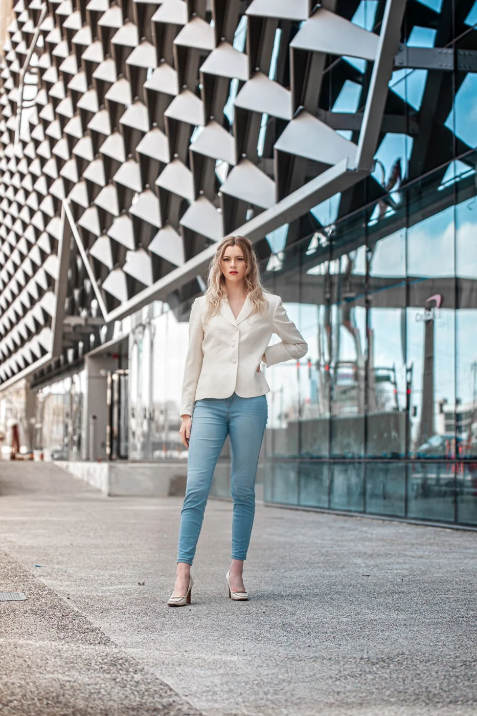 Frau in weißem Blazer, blauer Jeans und geschlossenen Schuhen vor einem Bürogebäude.