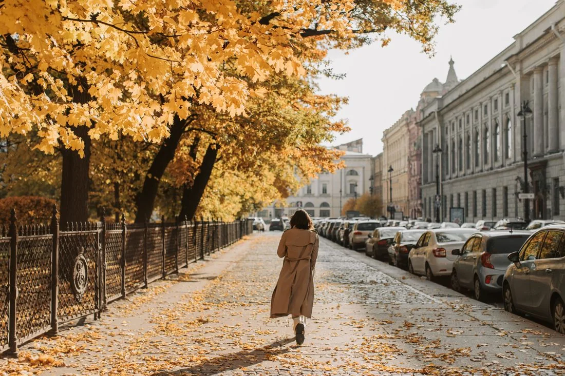 Frau im langen Trenchcoat auf einem herbstlichen Gehweg.