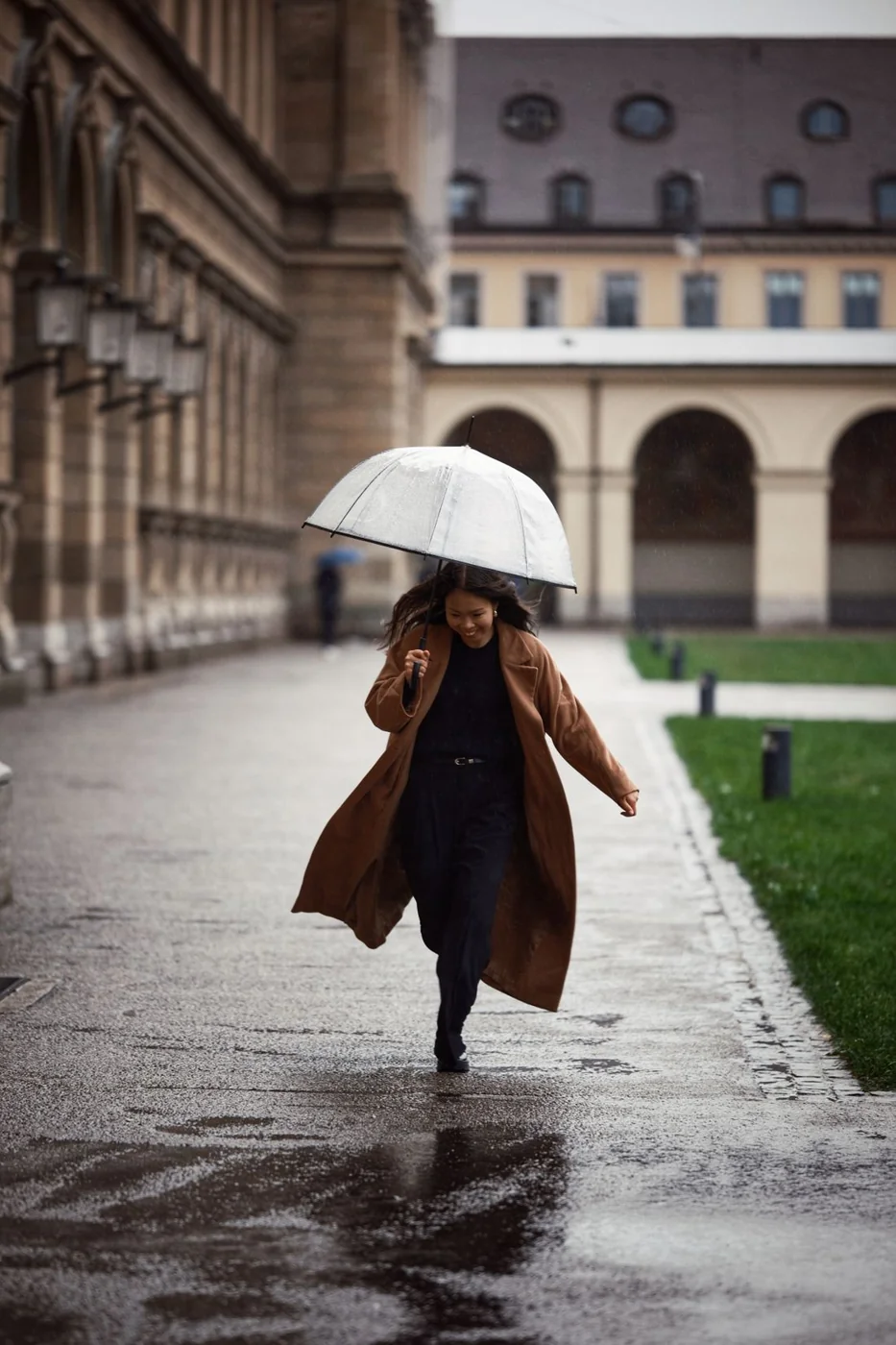 Frau in braunem Mantel geht mit einem Regenschirm durch die Stadt.